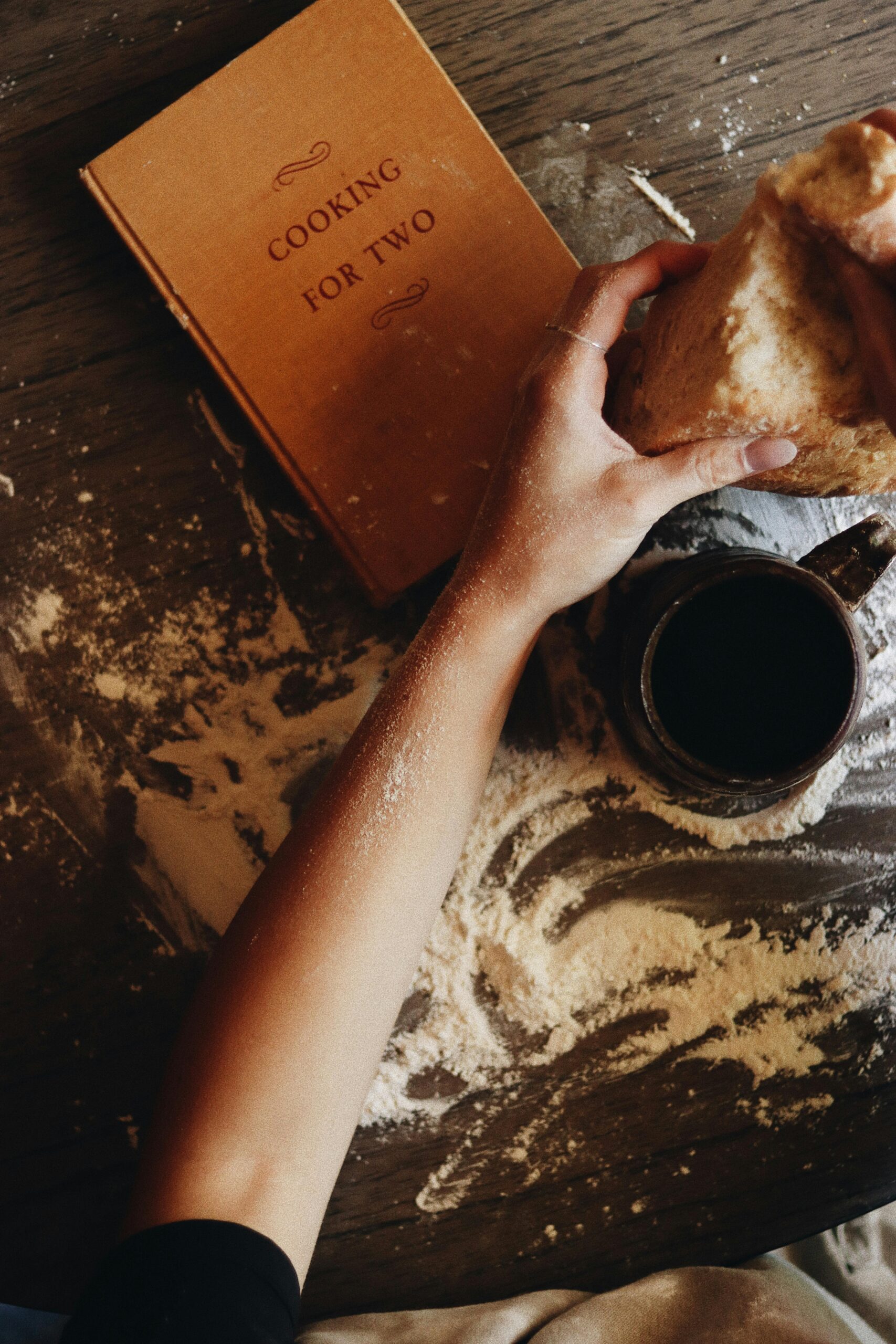Cozy kitchen scene with a hand holding bread next to coffee and a recipe book.